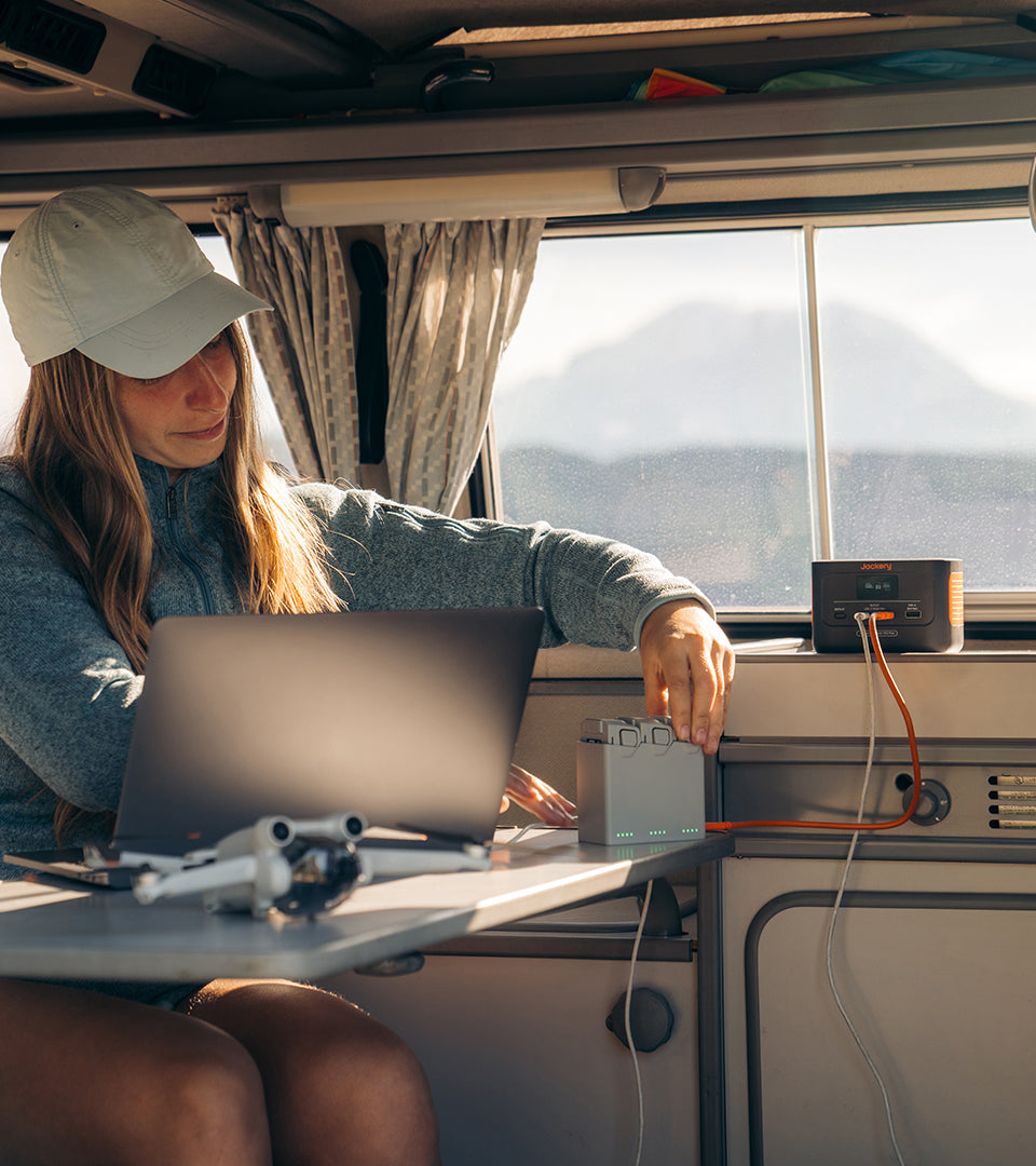 Inside a camper van, a person in a cap and long-sleeve top works on a laptop powered by the Jackery Explorer 100 Plus Portable Power Station. Its dependable LiFePO4 battery supports connected devices as blurred mountains appear outside the window.