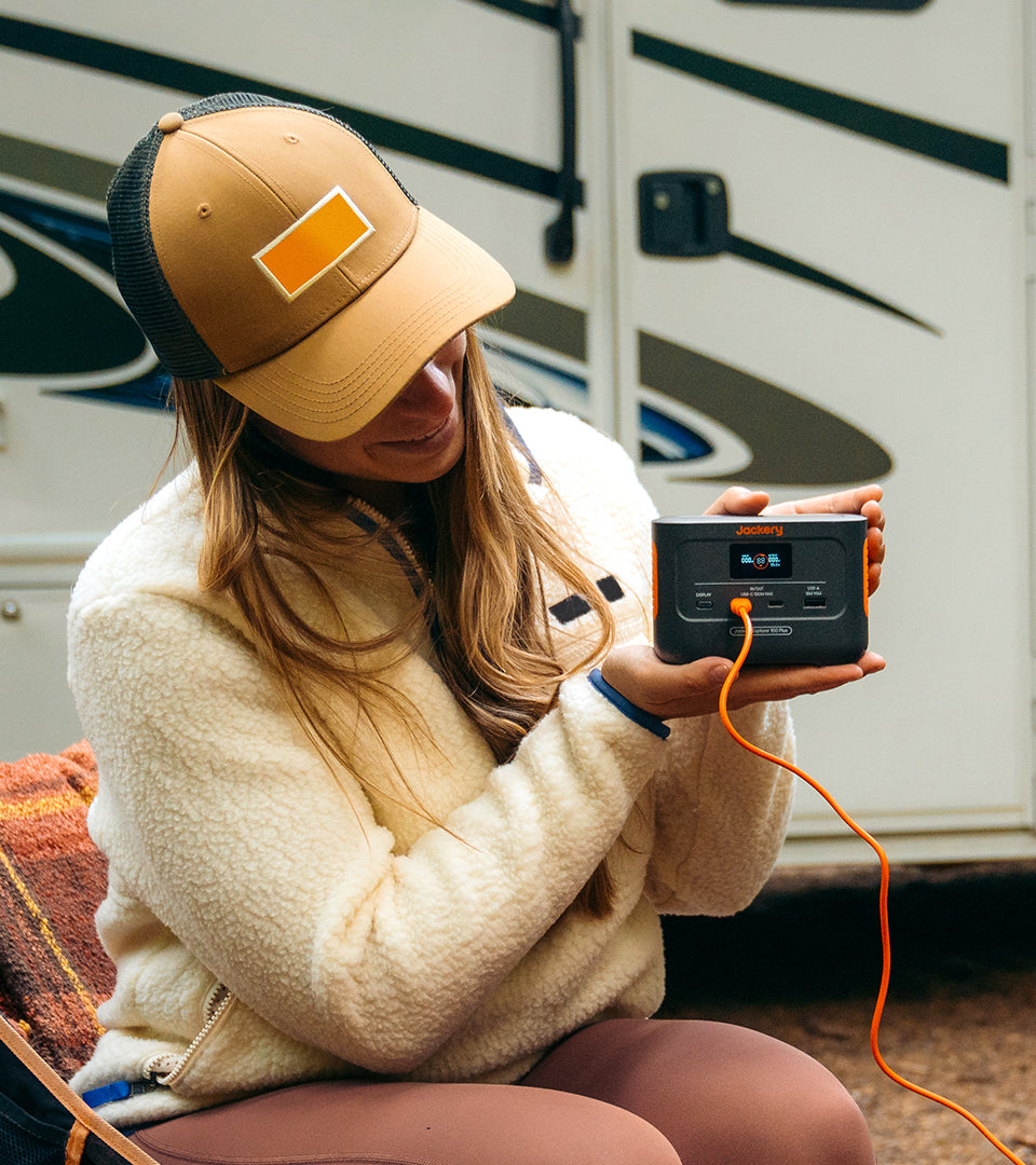 A person wearing a brown cap and light fleece jacket sits outdoors, holding a Jackery Explorer 100 Plus Portable Power Station with an orange cable. A vehicle door is visible in the background, hinting at camping or outdoor activities powered by its reliable LiFePO4 battery.