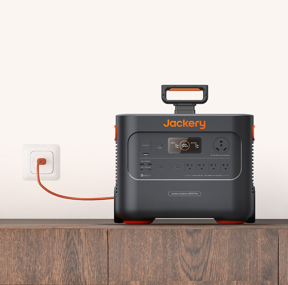 A Jackery Explorer 2000 Plus Portable Power Station rests on a wooden surface, connected to a wall outlet with a red cable. It features ultra-fast solar charging, a durable LiFePO4 battery, multiple ports, and a digital display for easy use.