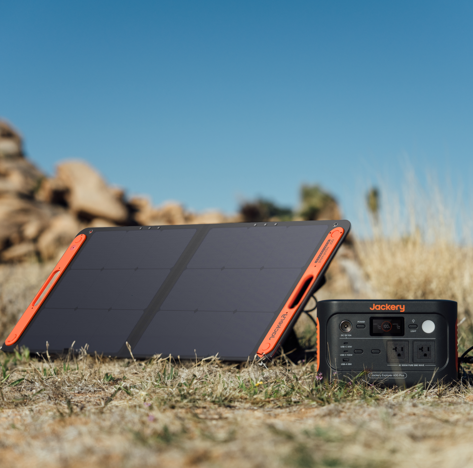 A portable solar panel charges the Jackery Explorer 600 Plus Portable Power Station outdoors on grass, showcasing its durability under a clear blue sky with rocky terrain in the background.