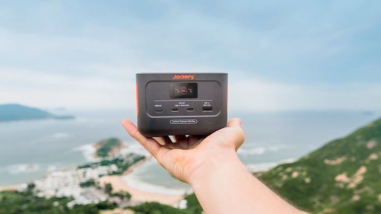 A person holds the Jackery Explorer 100 Plus Portable Power Station against a scenic ocean and hill backdrop. This gray device with orange accents includes multiple outlets, a display screen, and features a durable LiFePO4 battery.