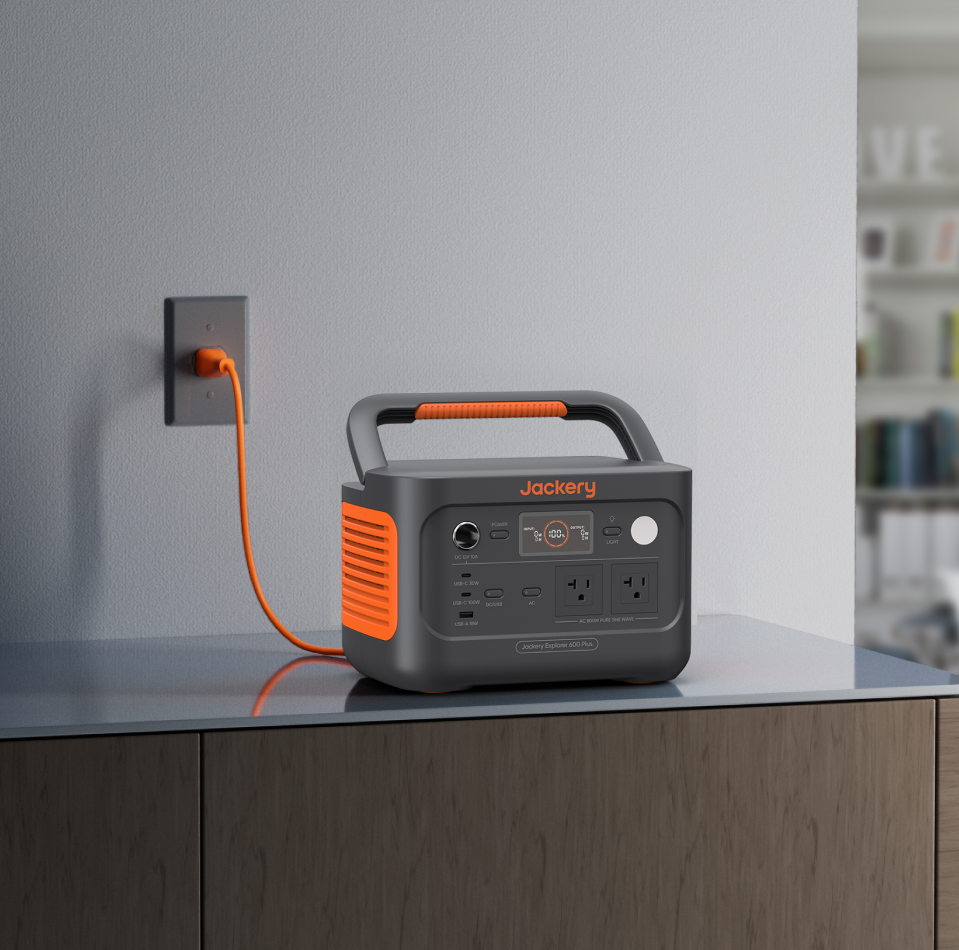 The Jackery Explorer 600 Plus Portable Power Station sits on a wooden countertop, plugged into a wall socket with an orange power cord. It boasts a handle and multiple front panel ports, showcasing its outdoor durability and adventure-ready versatility.