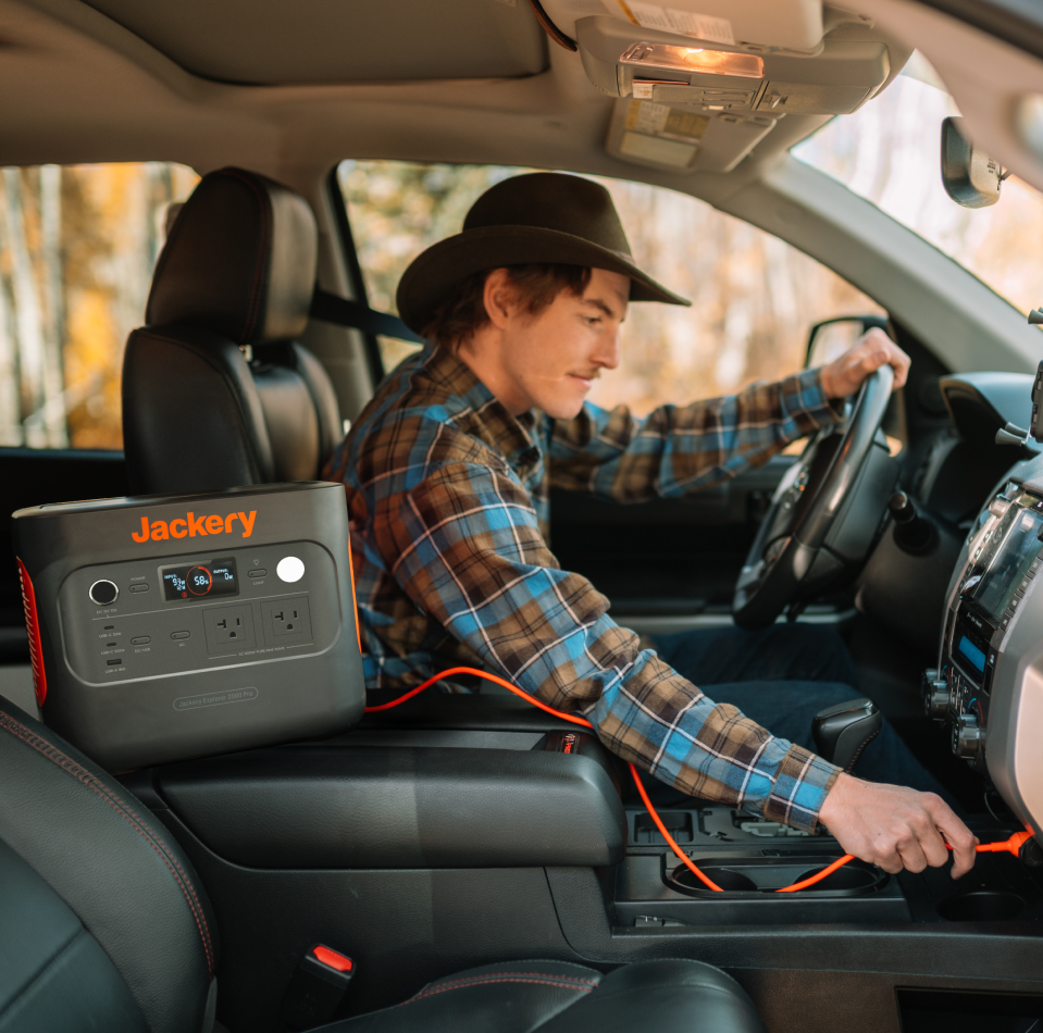 In a car, a person in a plaid shirt and hat adjusts the controls. The Jackery Explorer 600 Plus portable power station, known for its outdoor durability, sits beside them, connected with cables and ready to charge or power devices on the go.