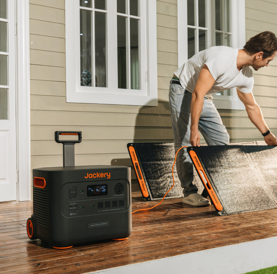 A person sets up a portable solar panel on a wooden deck next to a house, highlighting the Ultra Fast Solar Charging feature. Nearby, the Jackery Explorer 2000 Plus Portable Power Station with visible control panels complements the scene. They're in a white shirt and light gray pants beside white-framed windows.